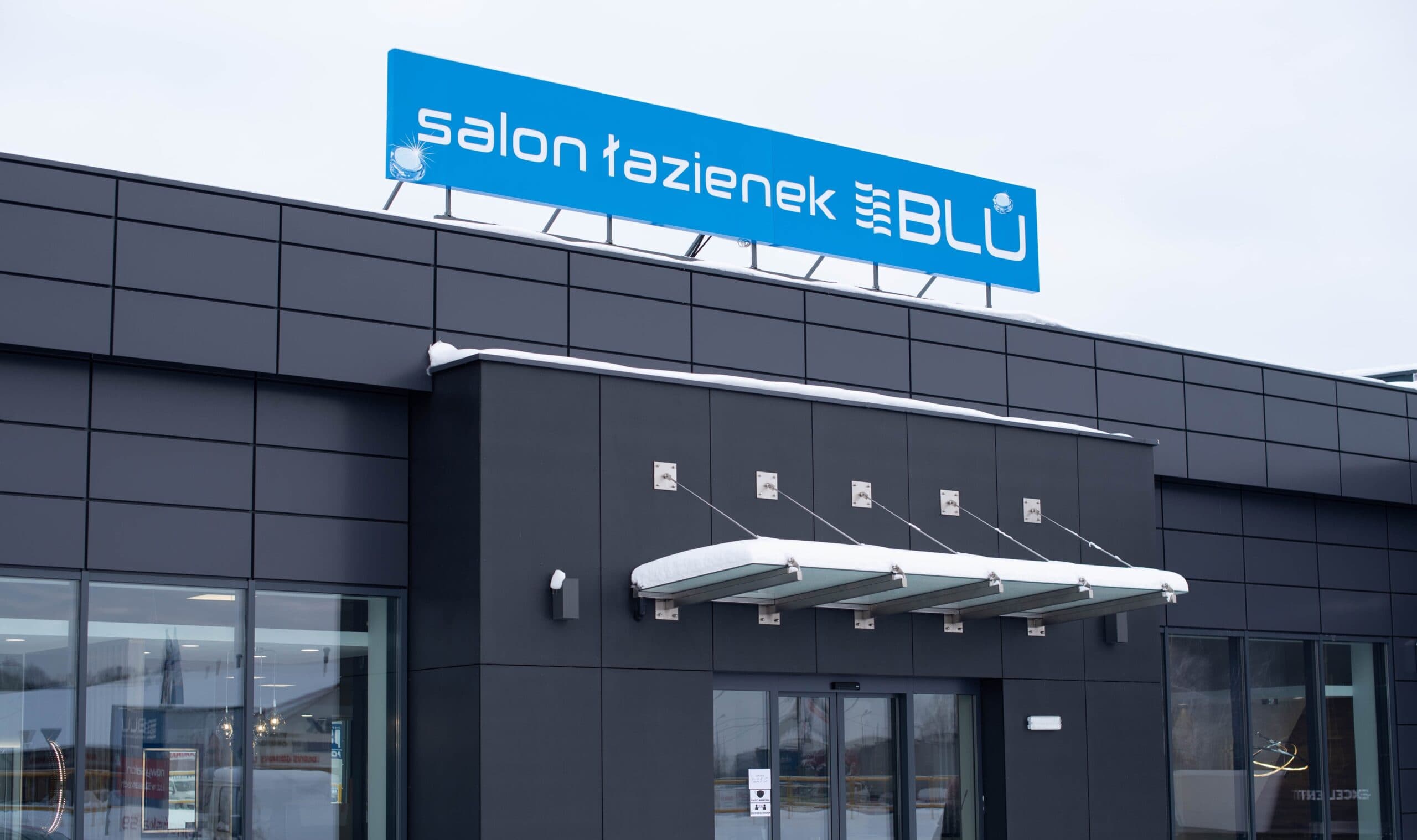 Elewacja budynku z niebieskim szyldem „salon łazienek BLU" nad wejściem, śnieg na daszku.