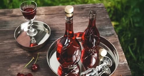 Dwie butelki i kieliszek z czerwonym napojem na metalowej tacy, obok wiśnie, w tle zieleń.