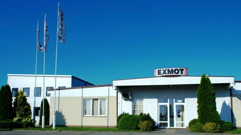 Budynek siedziby firmy Exmot z masztami flagowymi i szyldem z nazwą firmy.