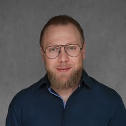 Mężczyzna z brodą i okularami w ciemnej koszuli na szarym tle.